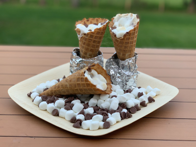 Two finished s'mores waffle cones standing up aluminum foil at the base, with mini marshmallows and chocolate chips and a third s'mores waffle cones on a white plate, sitting on a wood patio table, wi